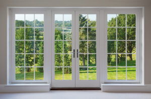 a set of finely crafted wooden Georgian style windows with doors set in a white frame, with white sills and caramel coloured walls. The view through the windows are towards a beautiful countryside garden.